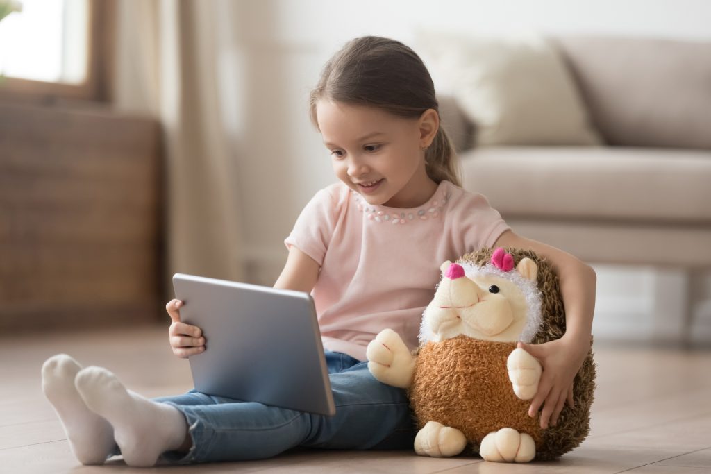 Image of Curious little child girl having fun using digital tablet alone embracing toy sitting on floor, happy preschool smart kid playing with computer looking at screen watching cartoons online at home